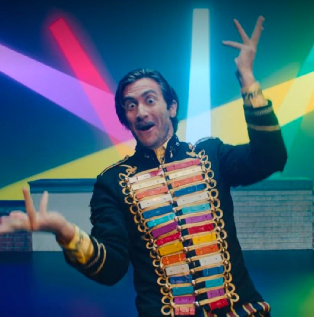Jake Gyllenhaal dancing, in front of rainbow lights, wearing an intricate blouse